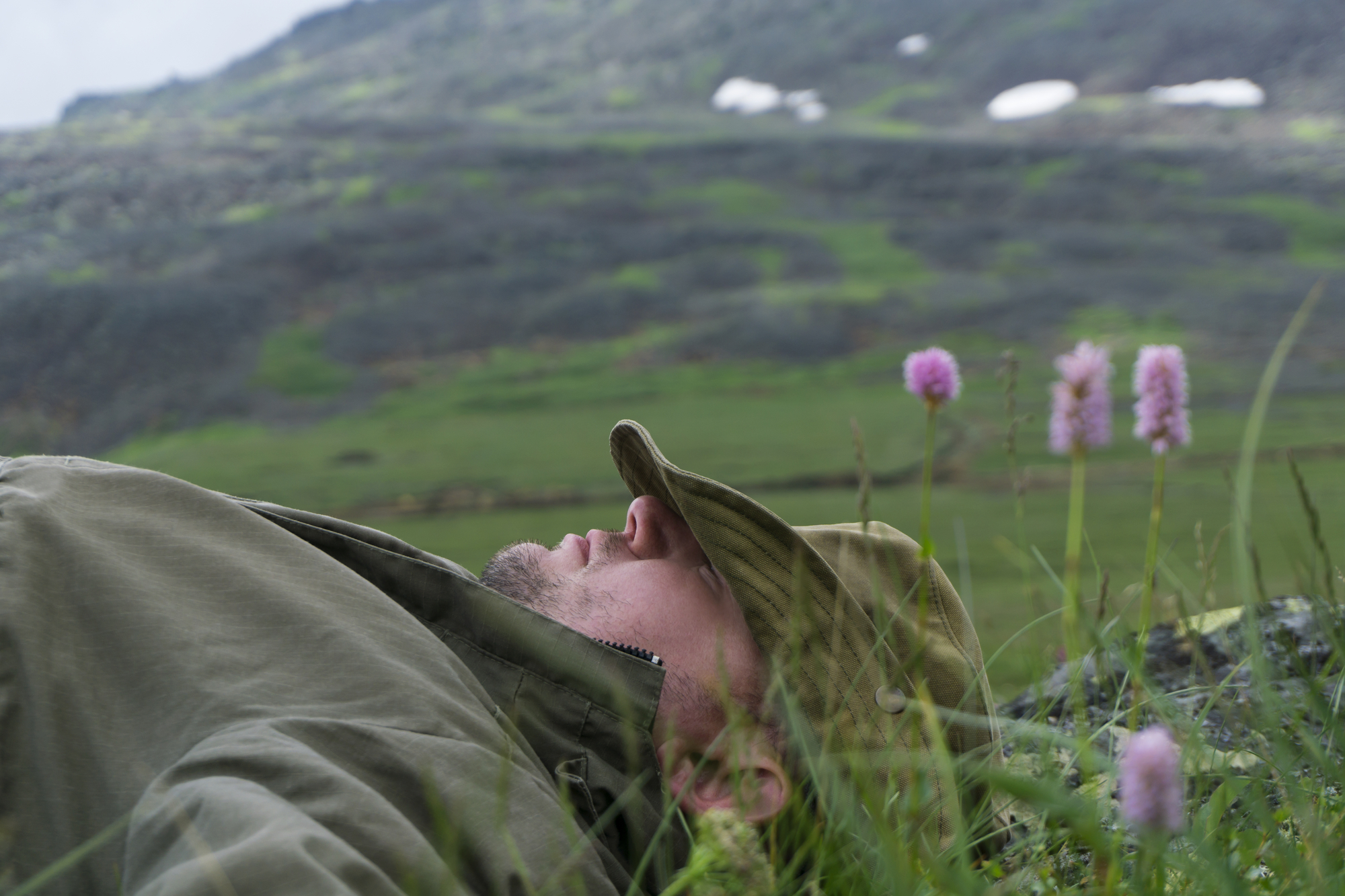 A person wearing a green jacket and hat lies on the grass in a mountainous area. The background features a blurred view of green hills with patches of snow, and pink wildflowers are in the foreground.