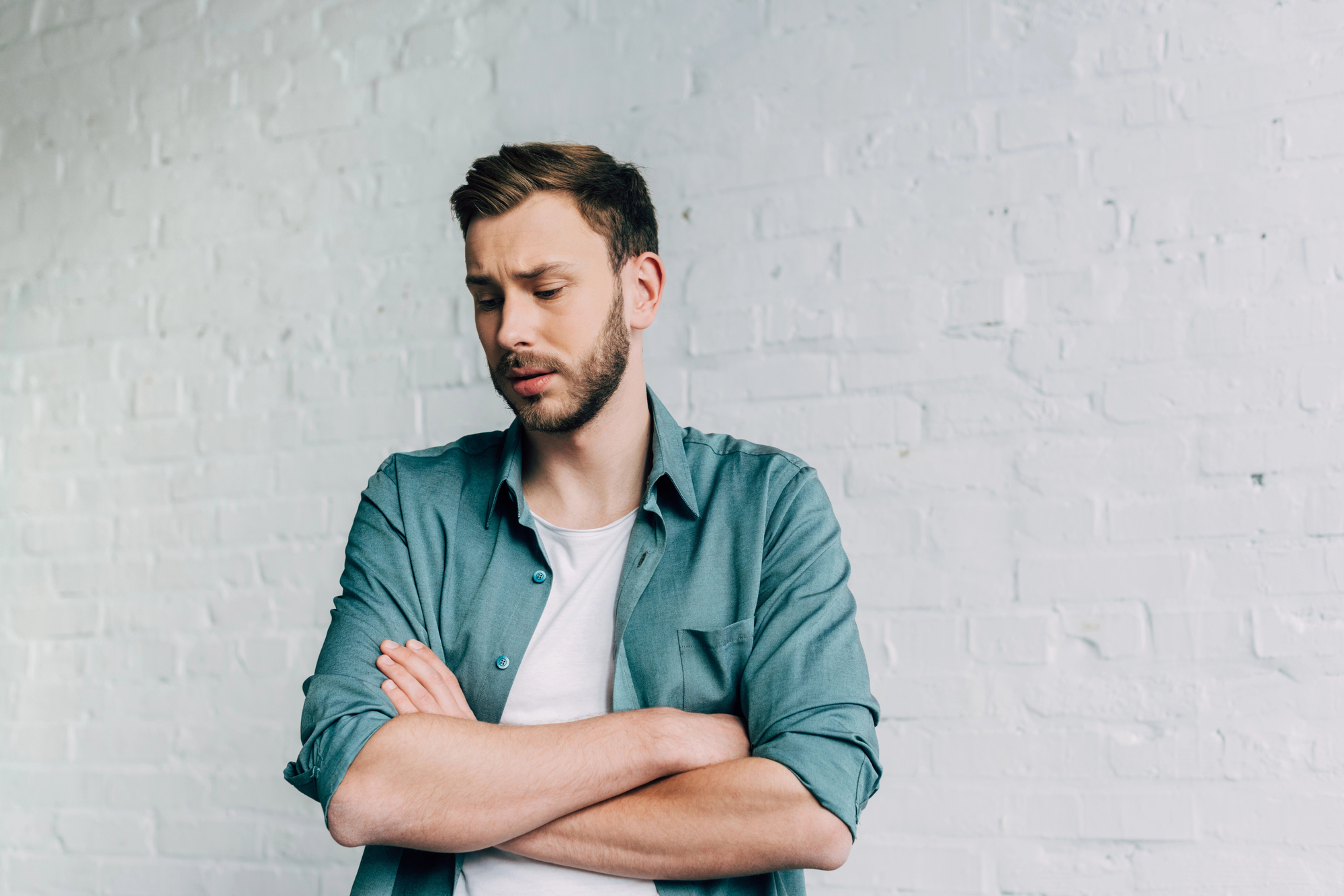 A man with short brown hair and a beard stands against a white brick wall, looking down with a concerned expression. He is wearing a green button-up shirt over a white t-shirt and has his arms crossed over his chest.