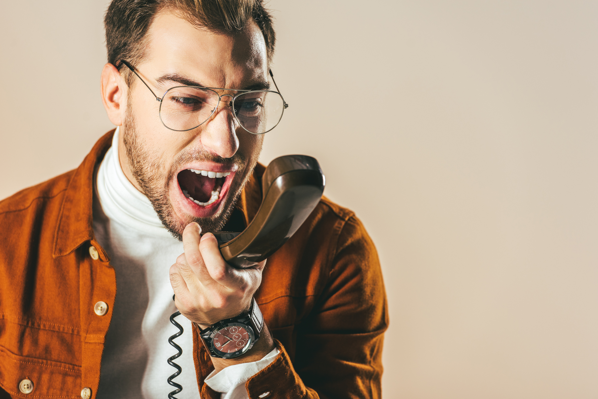 A man with glasses and a beard is angrily yelling into a phone receiver. He is wearing a white turtleneck and a brown jacket. The background is plain and neutral.