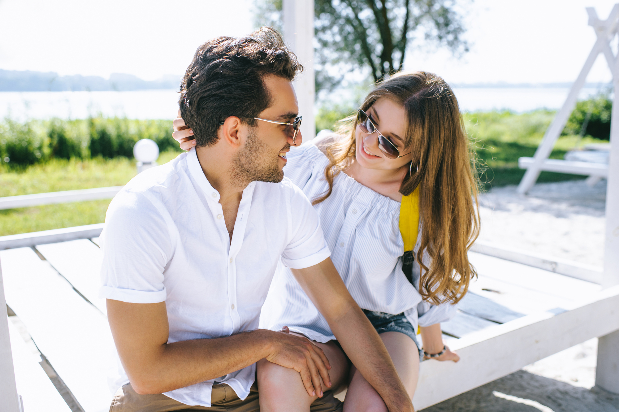 A man and woman sit together on a white bench outdoors on a sunny day. Both are wearing sunglasses and casual summer clothes. The woman, with long hair, smiles at the man while resting her hand on his shoulder. The background features greenery and a body of water.