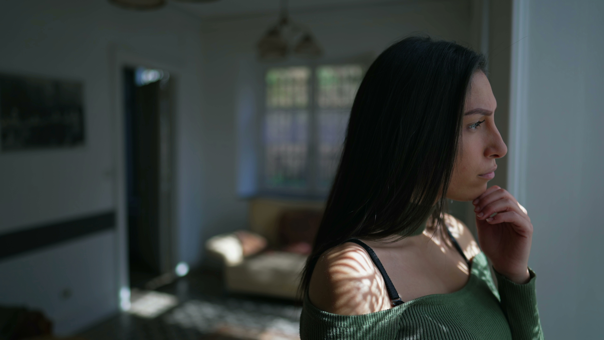 A woman with long dark hair stands indoors near a window, gazing contemplatively outside. Light filters through the window, casting shadows on her face. She wears a green off-the-shoulder top, and the background shows a blurred room with a couch and a window.