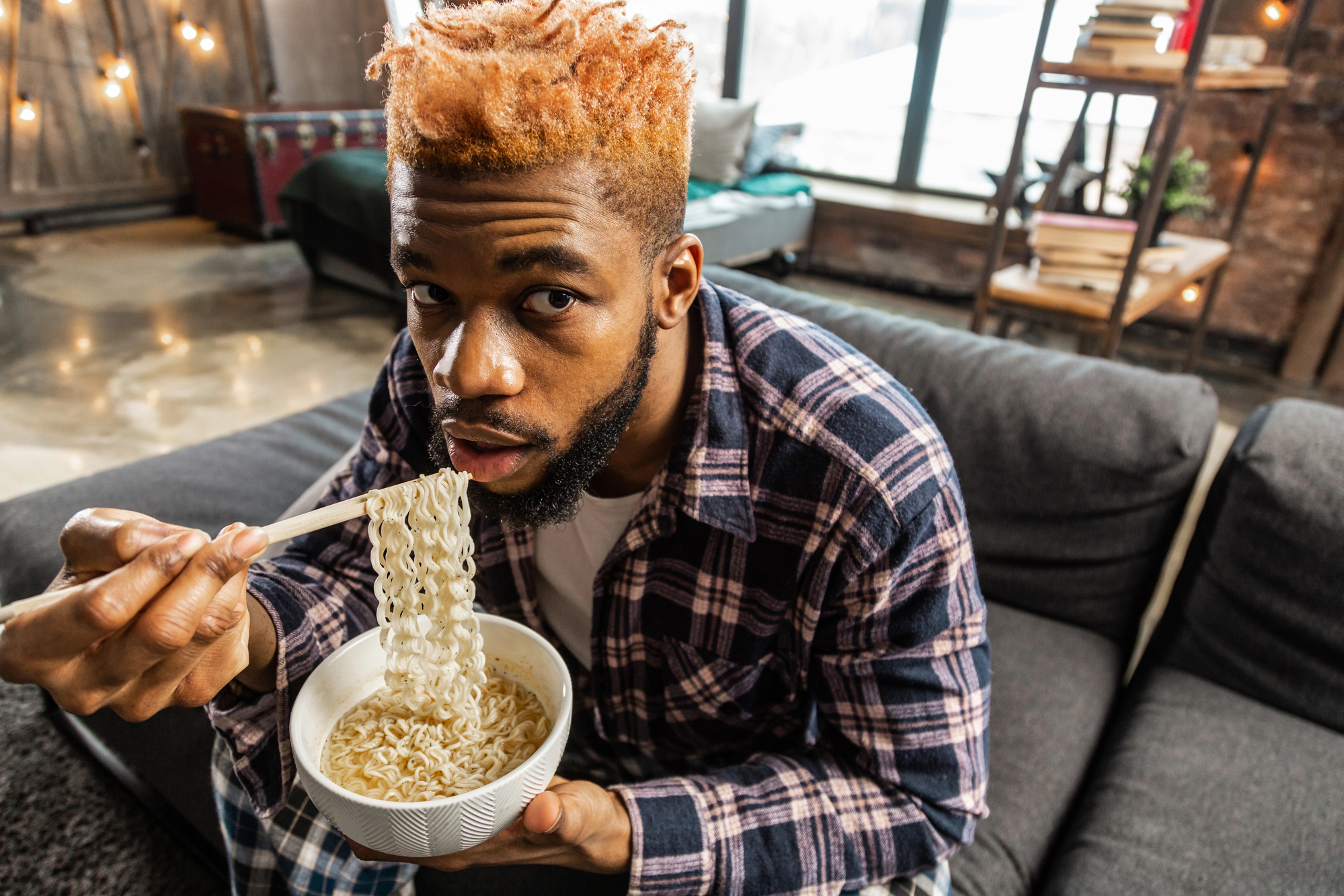 A man with orange-dyed hair, wearing a plaid shirt, sits on a sofa in a cozy, modern living room and eats noodles from a bowl using chopsticks. The living room features warm lighting, a wooden floor, and shelves with books and decorations.