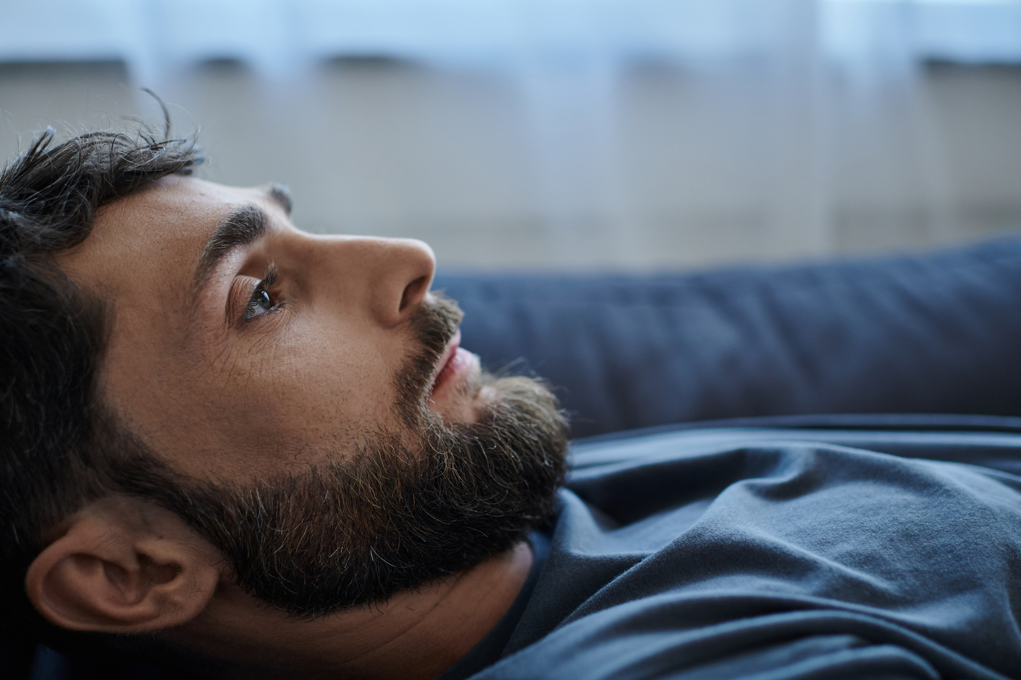 A close-up of a bearded man lying down on a couch, looking thoughtfully into the distance. He has short dark hair and is wearing a dark blue shirt. The background is softly blurred, highlighting his profile and contemplative expression.