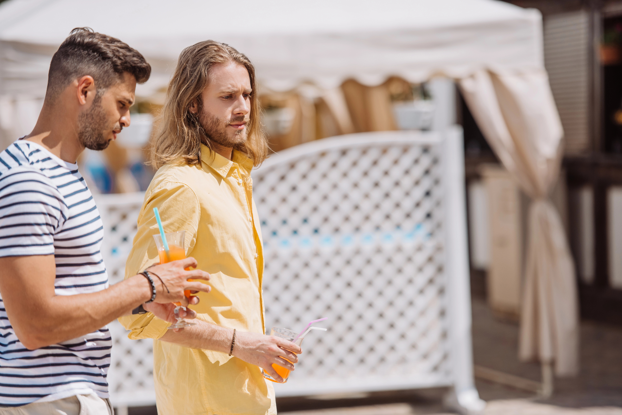 Two men are walking outdoors, each holding a drink with a straw. One is wearing a striped shirt, and the other is wearing a yellow shirt. They appear to be engaged in conversation. A white lattice and a canopy are visible in the background.