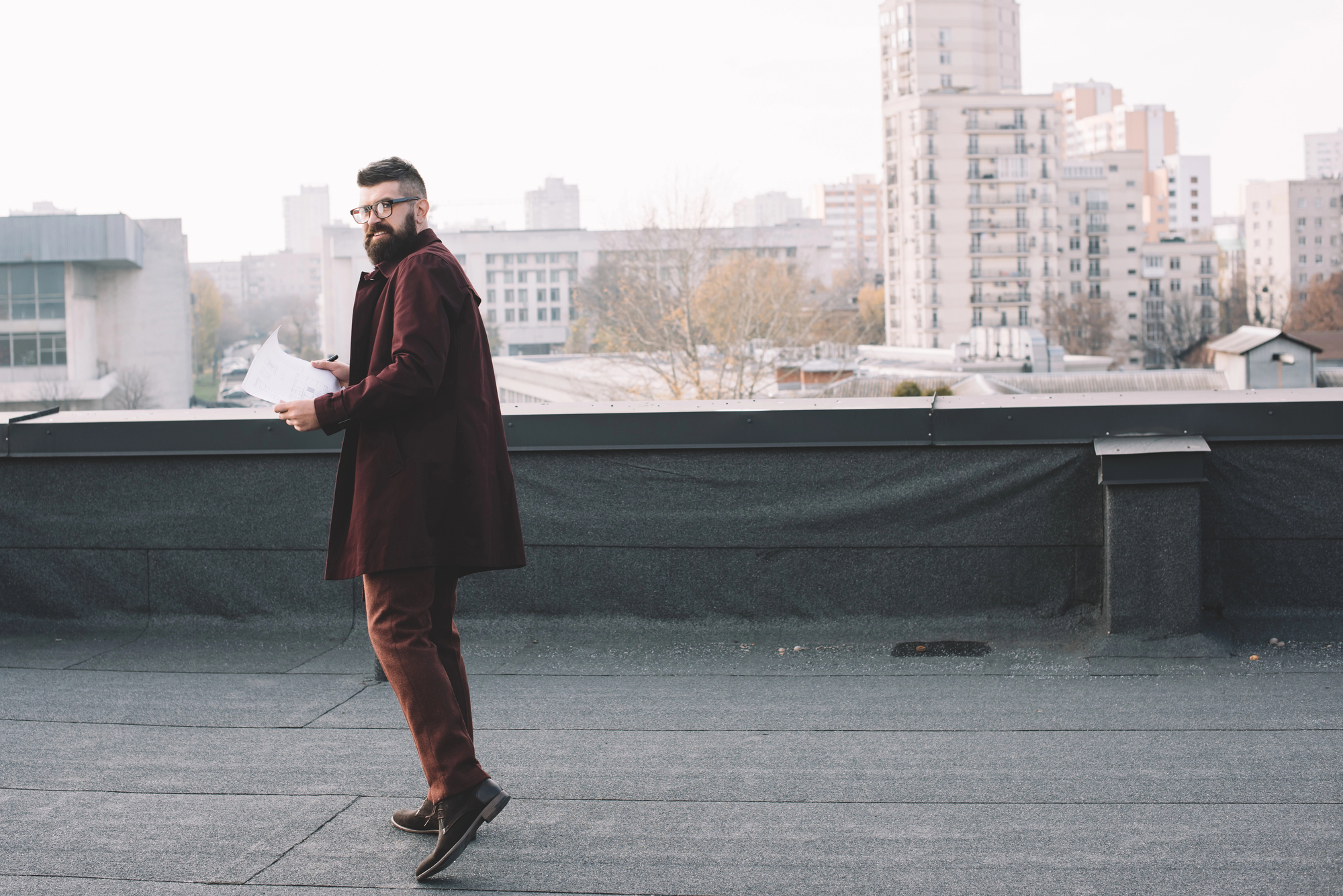 A bearded man in glasses and a maroon coat is holding papers and walking on a rooftop. Urban buildings and structures are visible in the background on a hazy day.