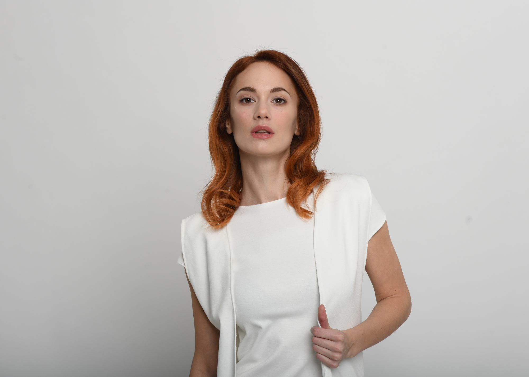 A woman with shoulder-length red hair is standing against a plain white background. She is wearing a white outfit and gazing directly at the camera with a neutral expression. One hand is slightly lifting the edge of her top.