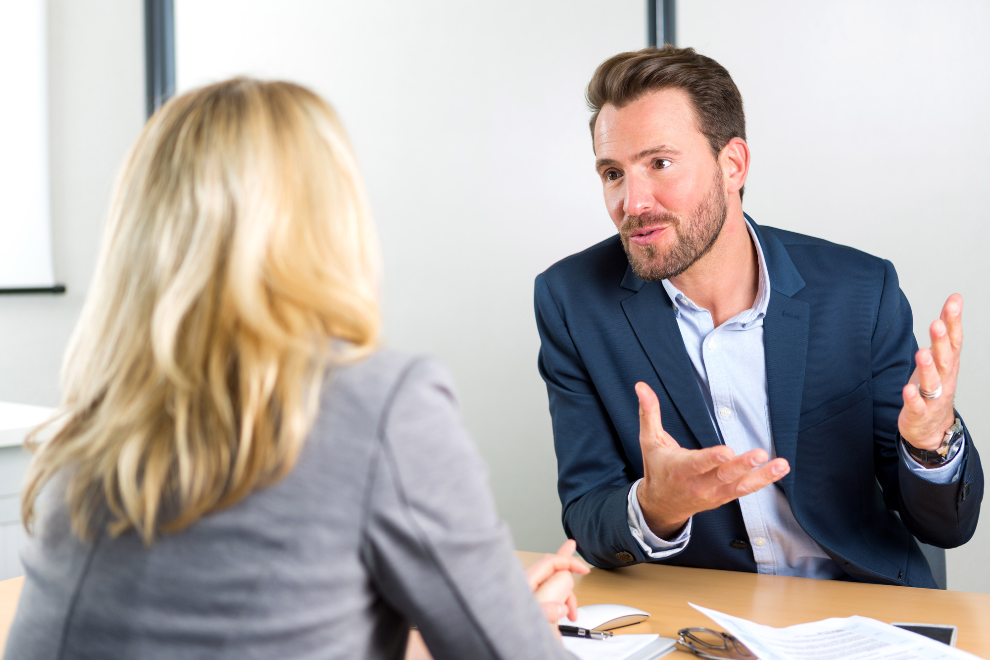 Two people are having a discussion at a desk in a professional setting. The person on the right, wearing a blue blazer, is speaking animatedly with hands gesturing, while the person on the left, with blonde hair in a gray blazer, is listening attentively.