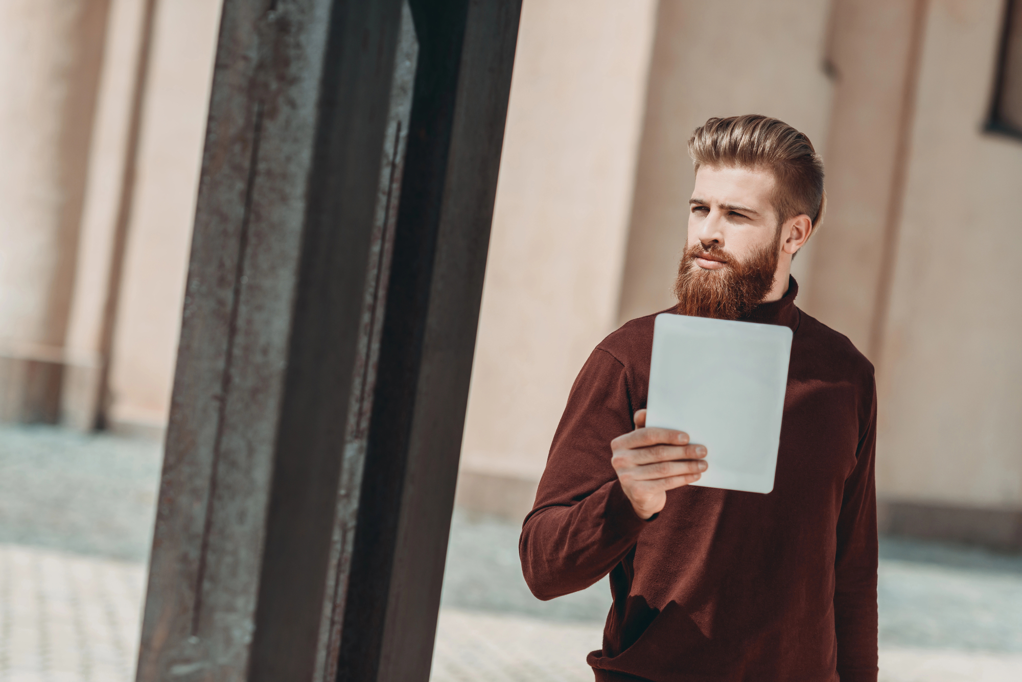A bearded man with styled hair wearing a maroon sweatshirt is standing outside holding a tablet. He gazes to the right with a thoughtful expression. The background shows a blurred building and cobblestone ground.