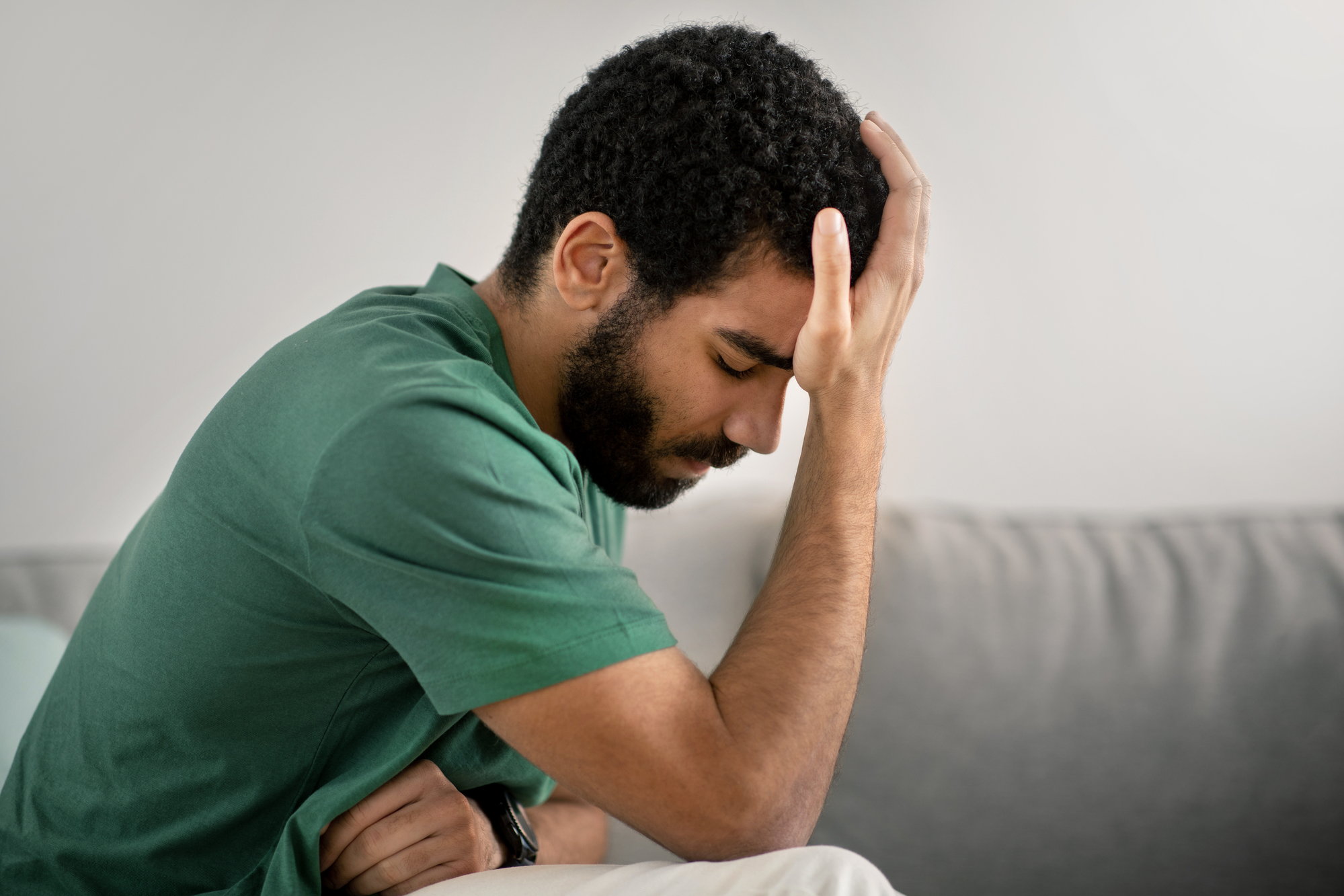A man with short curly hair and a beard sits on a couch, resting his elbow on his knee and his head in his hand. He is wearing a green t-shirt and appears to be deep in thought or stressed, with a worried expression on his face.