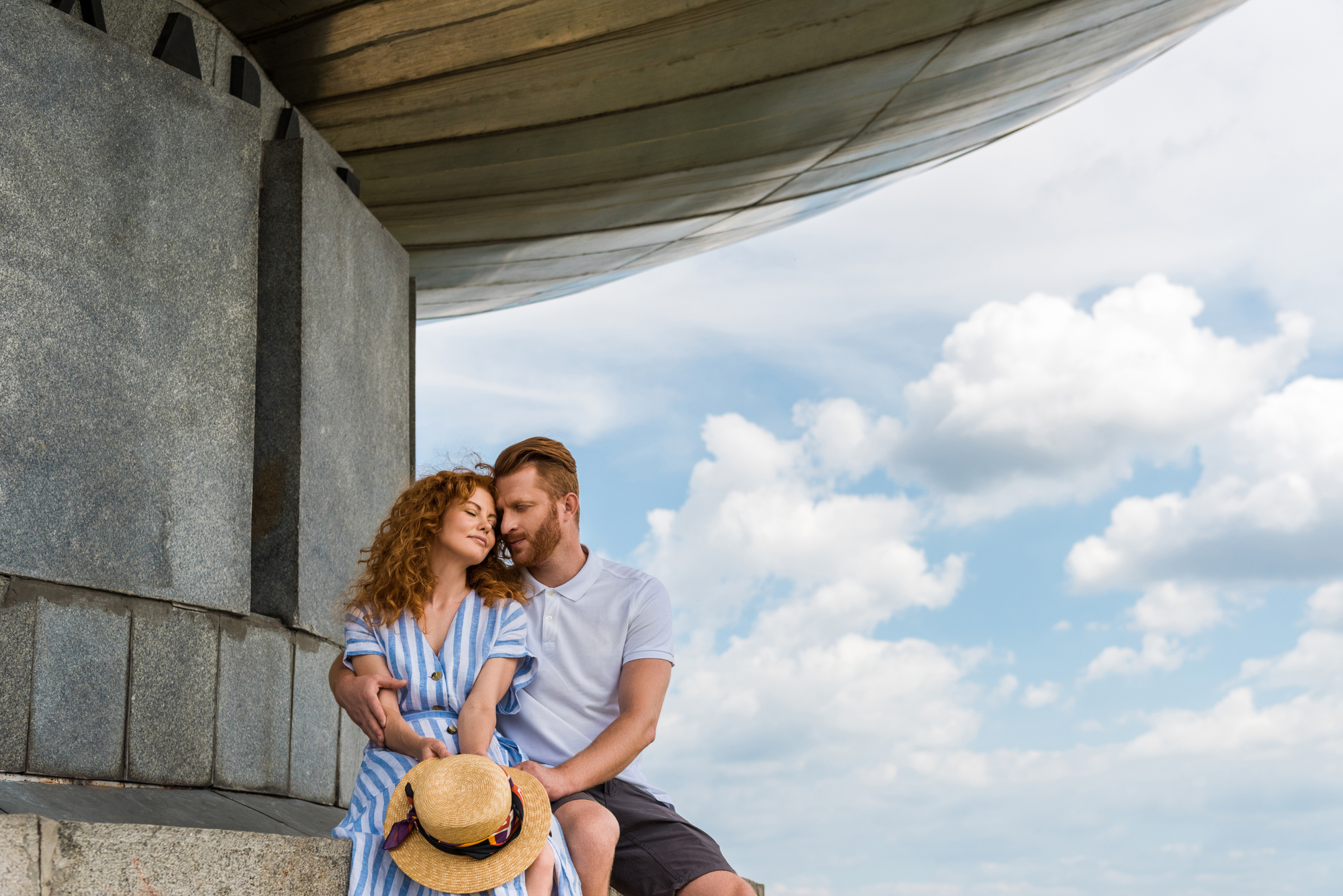 A couple sits closely together under a large, modern metallic sculpture. The woman in a blue striped dress holds a straw hat and rests her head on the man's shoulder, who is in a white shirt and shorts. The sky is partly cloudy, creating a serene backdrop.