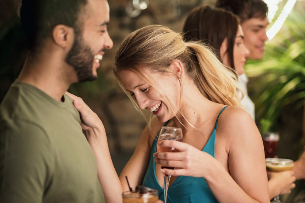 A group of young adults socializing at a casual indoor gathering. A woman in the foreground, holding a drink, is laughing while interacting with a smiling man. Other people are engaged in conversation in the background. The atmosphere appears lively and cheerful.