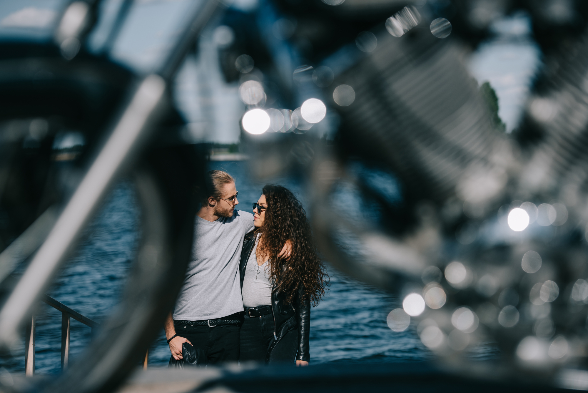 A man and woman stand by a waterfront, arm in arm, smiling at each other. The scene is partially obstructed by the out-of-focus frame and engine of a motorcycle in the foreground, creating a sense of depth and context.