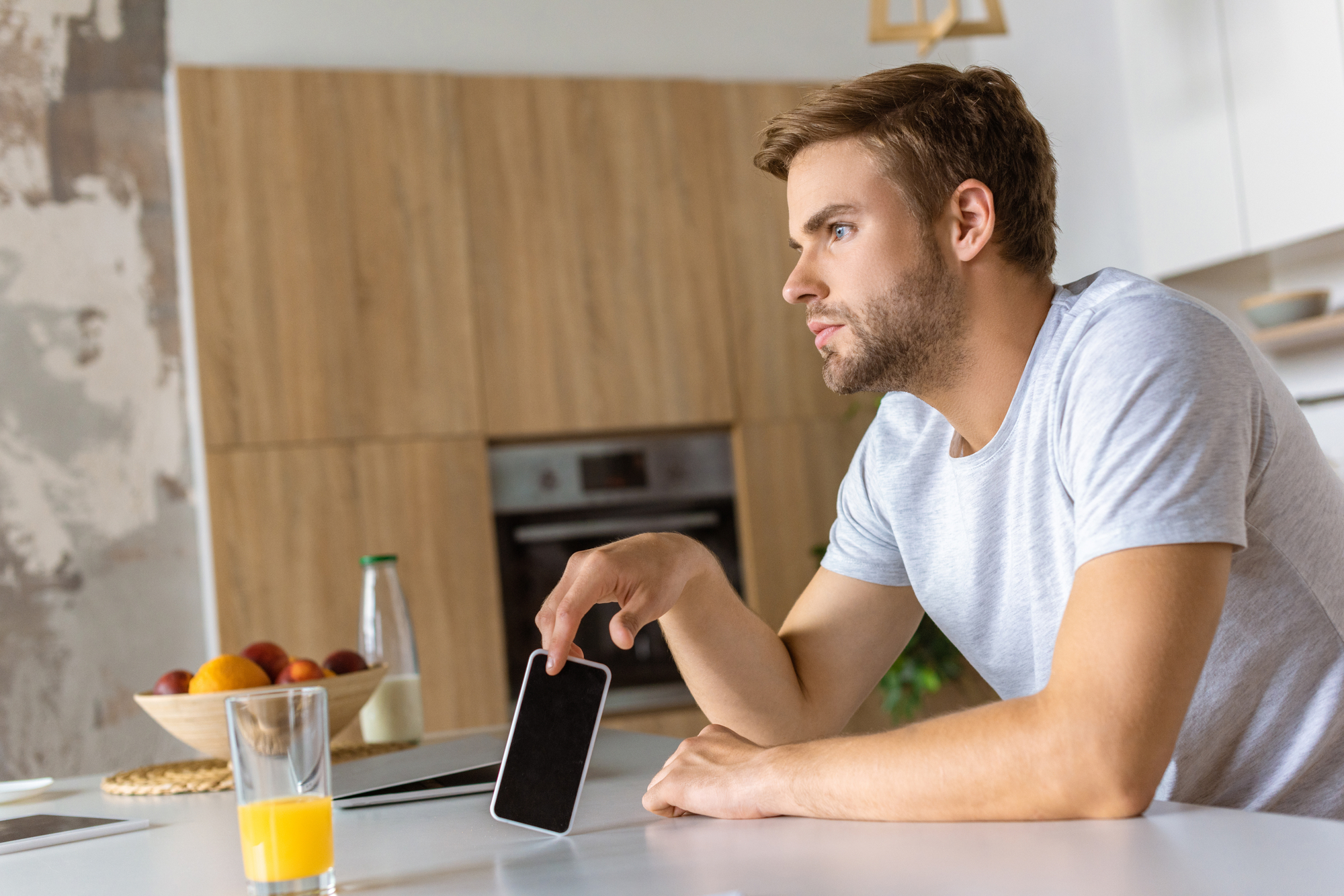 A bearded man with short hair wearing a gray t-shirt sits at a white kitchen table, looking pensively into the distance while holding a smartphone. A glass of orange juice and a bowl of fruit are on the table, with wooden cabinets and a textured wall in the background.