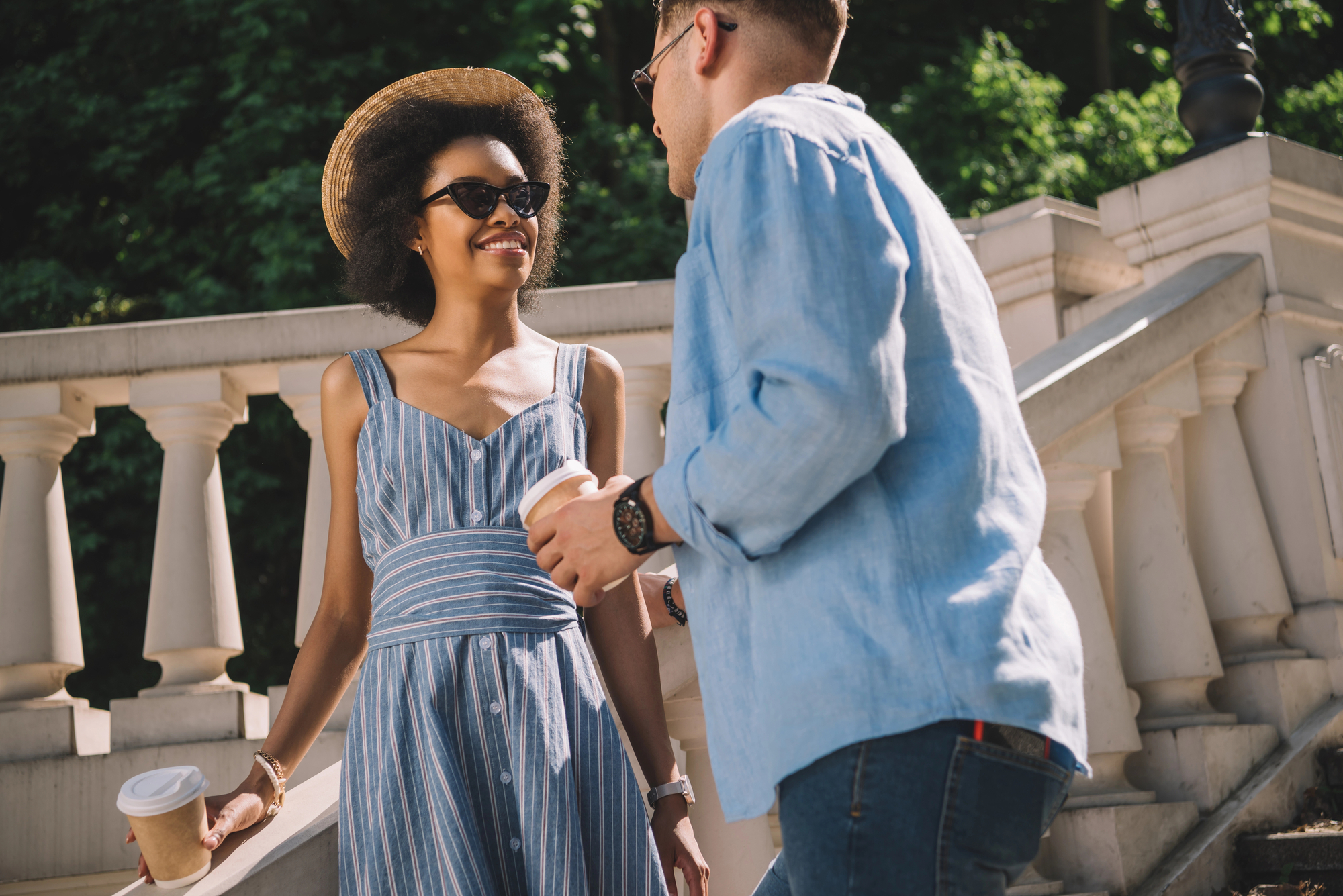 Two people are outdoors near a staircase, holding coffee cups. The person on the left is wearing a striped dress, sunglasses, and a hat, and is smiling. The person on the right is wearing a light blue shirt and eyeglasses, and appears to be talking. Trees are in the background.
