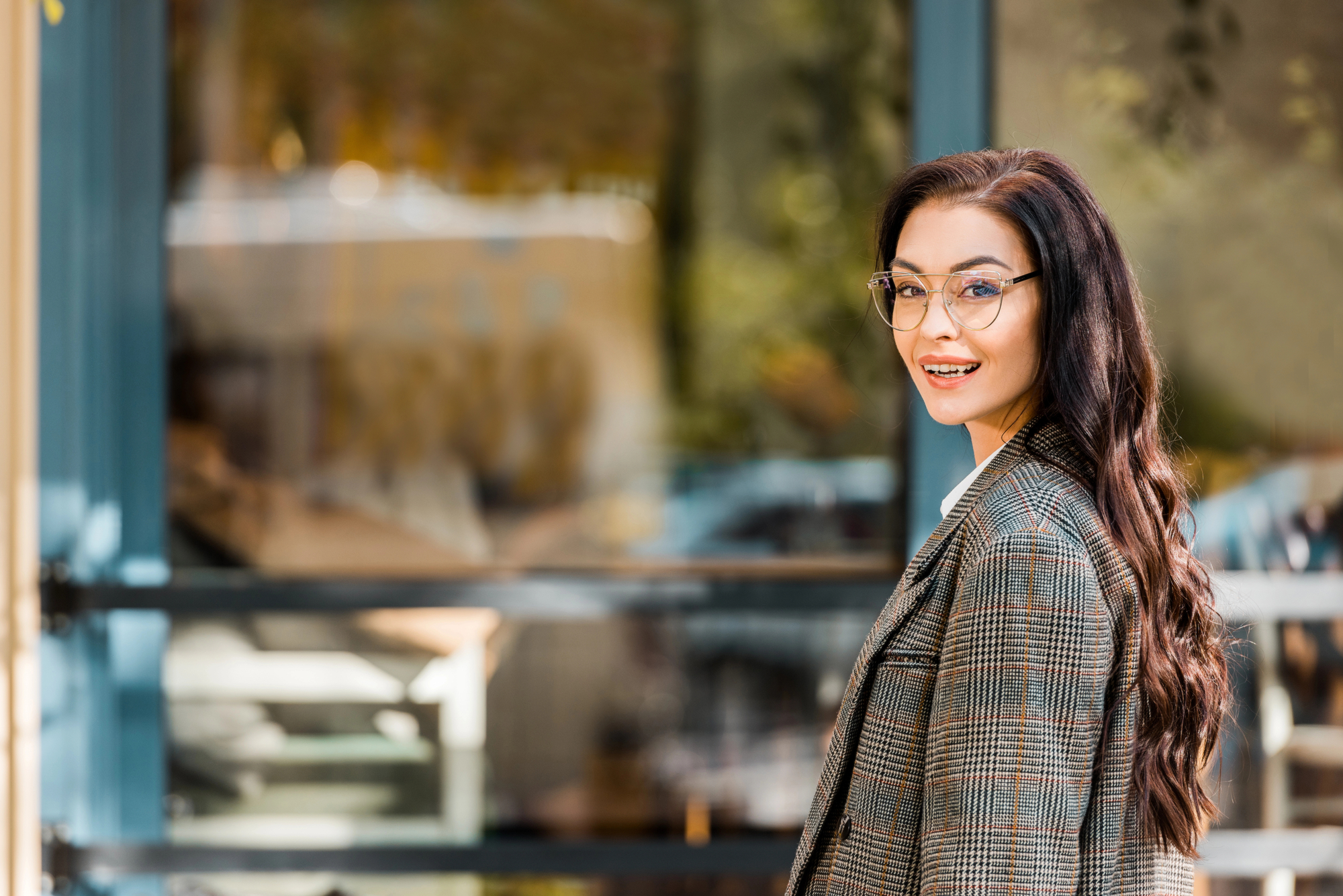 A woman with long, dark hair and glasses, dressed in a plaid coat, smiles while standing outside near a blue-framed glass window. The background shows a blurred view of trees and buildings. She is turning slightly towards the camera.
