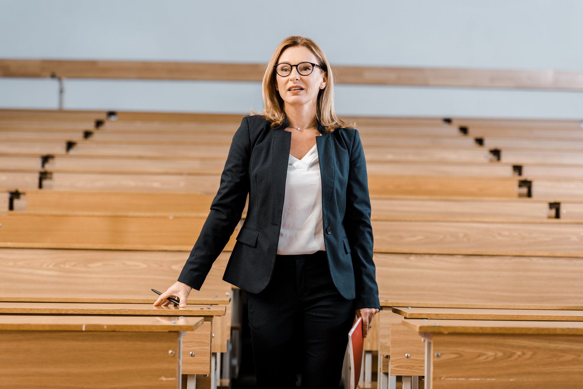A woman in a black suit and white blouse stands confidently in an empty lecture hall with wooden benches, smiling softly and holding a folder and pen. She has shoulder-length blond hair and wears glasses.