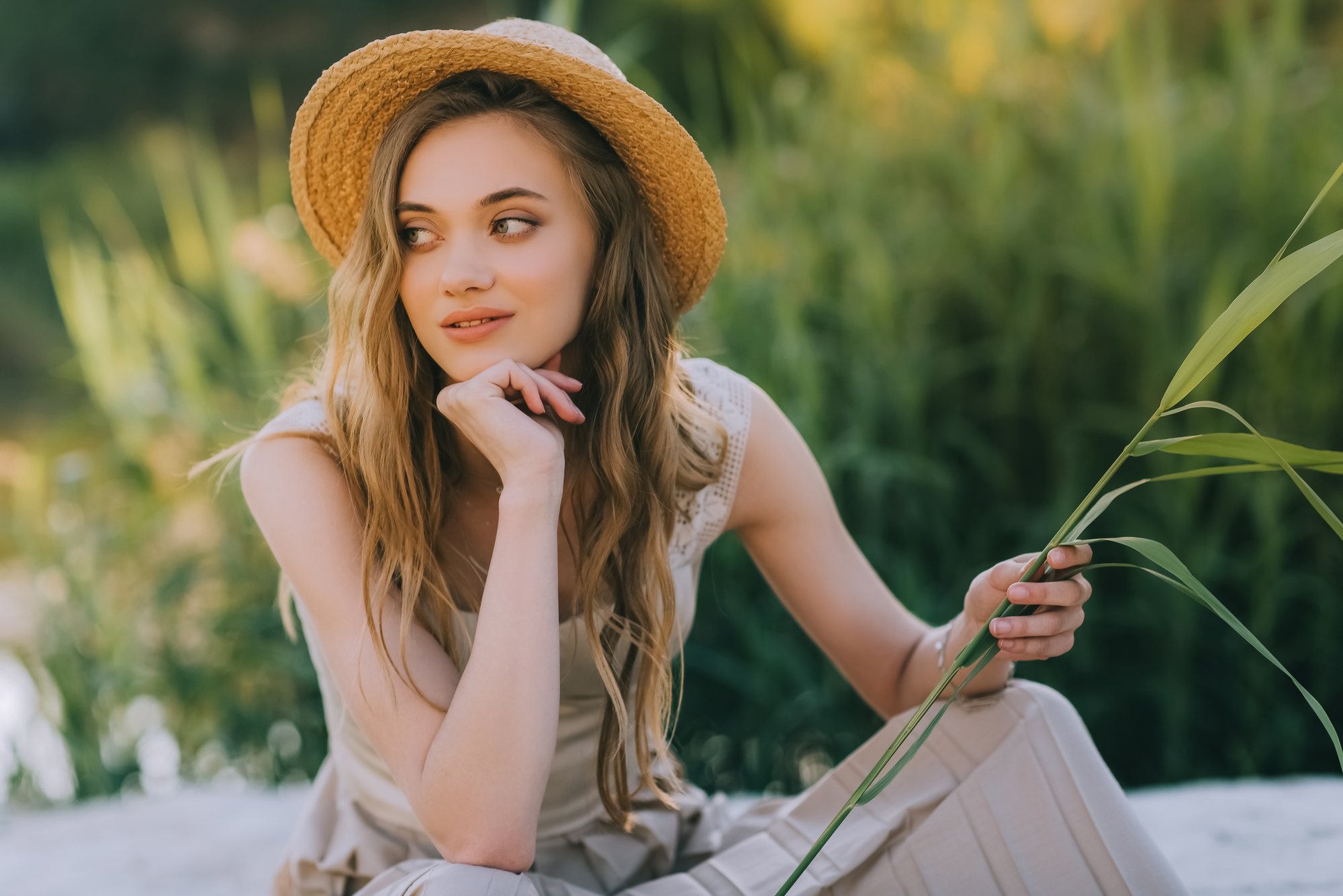 A young woman with long, wavy hair sits outdoors on a sunny day, wearing a straw hat and a beige outfit. She gazes thoughtfully to the side, resting her chin on her hand, and holds a long blade of grass in her other hand. Green foliage is visible in the background.