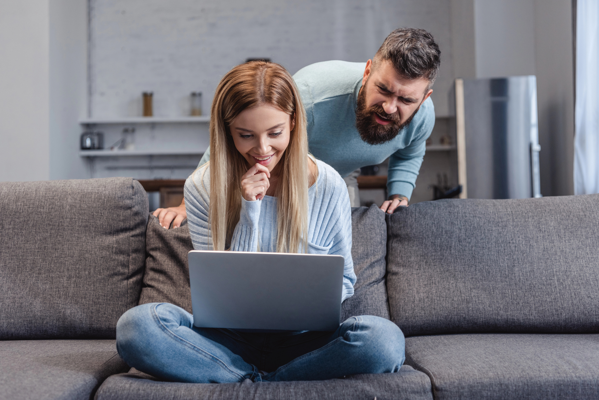 A woman sits on a grey sofa with her laptop on her lap, looking at the screen and smiling. A man stands behind the sofa, leaning forward with a curious expression, looking at her screen. They are in a modern living room with a minimalistic decor.