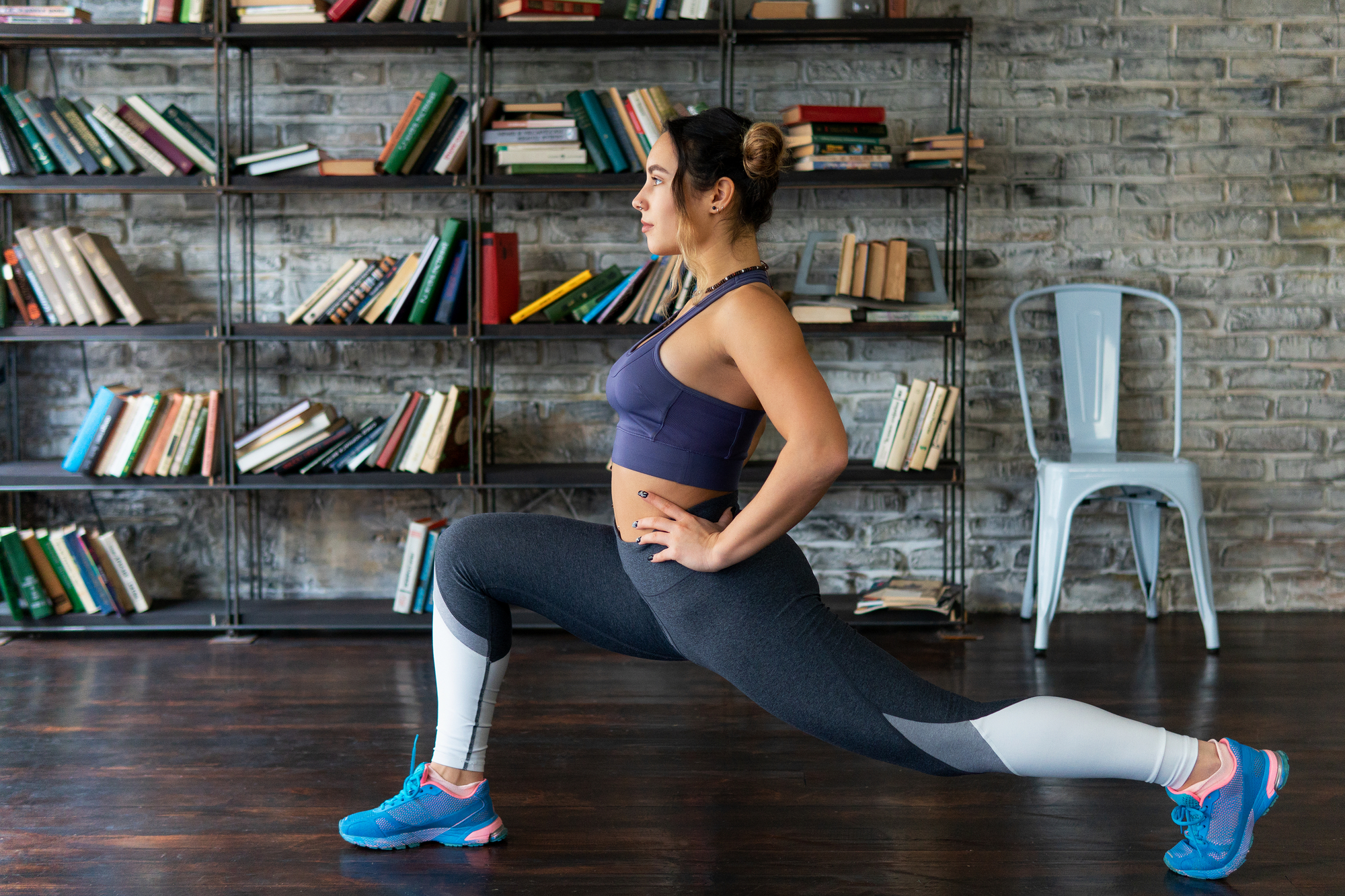 A woman in athletic wear performs a lunge exercise in front of a bookshelf with scattered books. She wears a blue sports bra, gray leggings, and blue athletic shoes. A metal chair is positioned against the brick wall background.