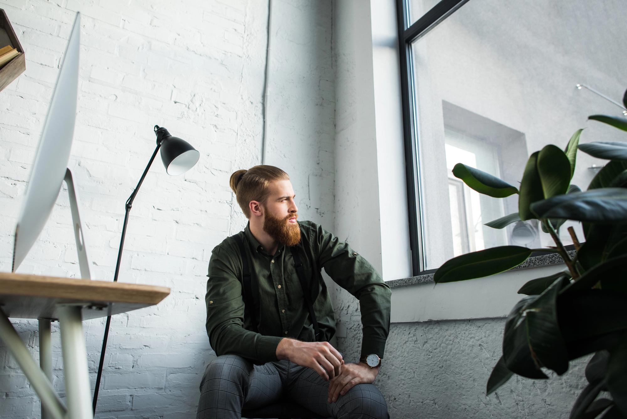 A bearded man with a top knot hairstyle is sitting by a large window in a modern office, gazing outside. He is wearing a dark green shirt and checkered pants. A desk with a computer monitor and a black lamp are on his left, and a large plant is on his right.