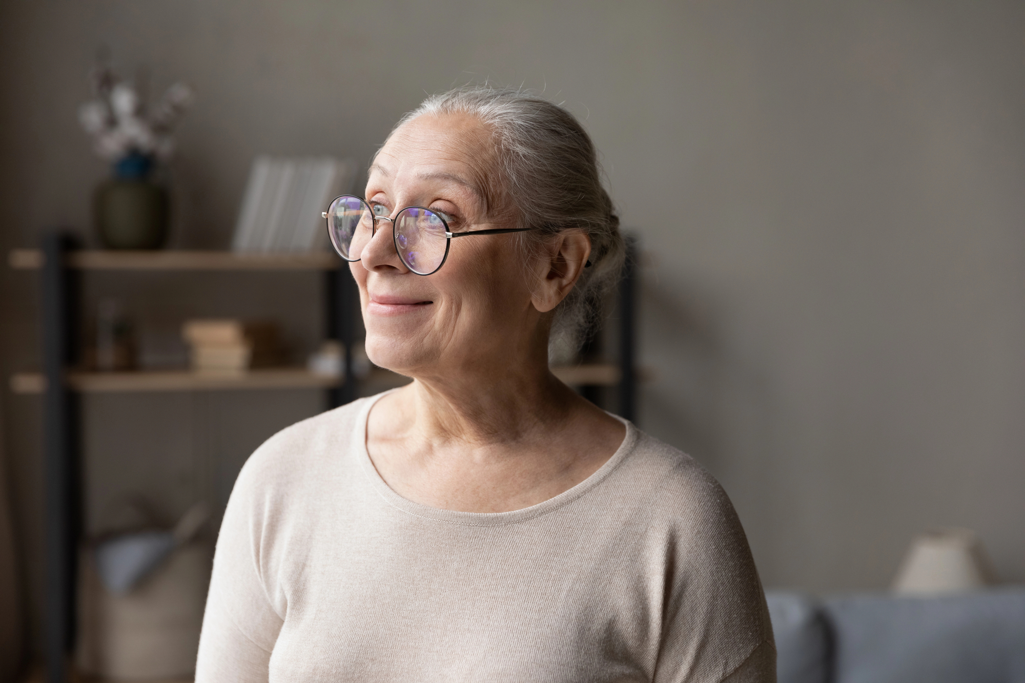 An elderly woman with gray hair tied back is wearing round glasses and a light beige sweater. She is smiling and looking to the side in a cozy room with a shelf and soft lighting in the background.