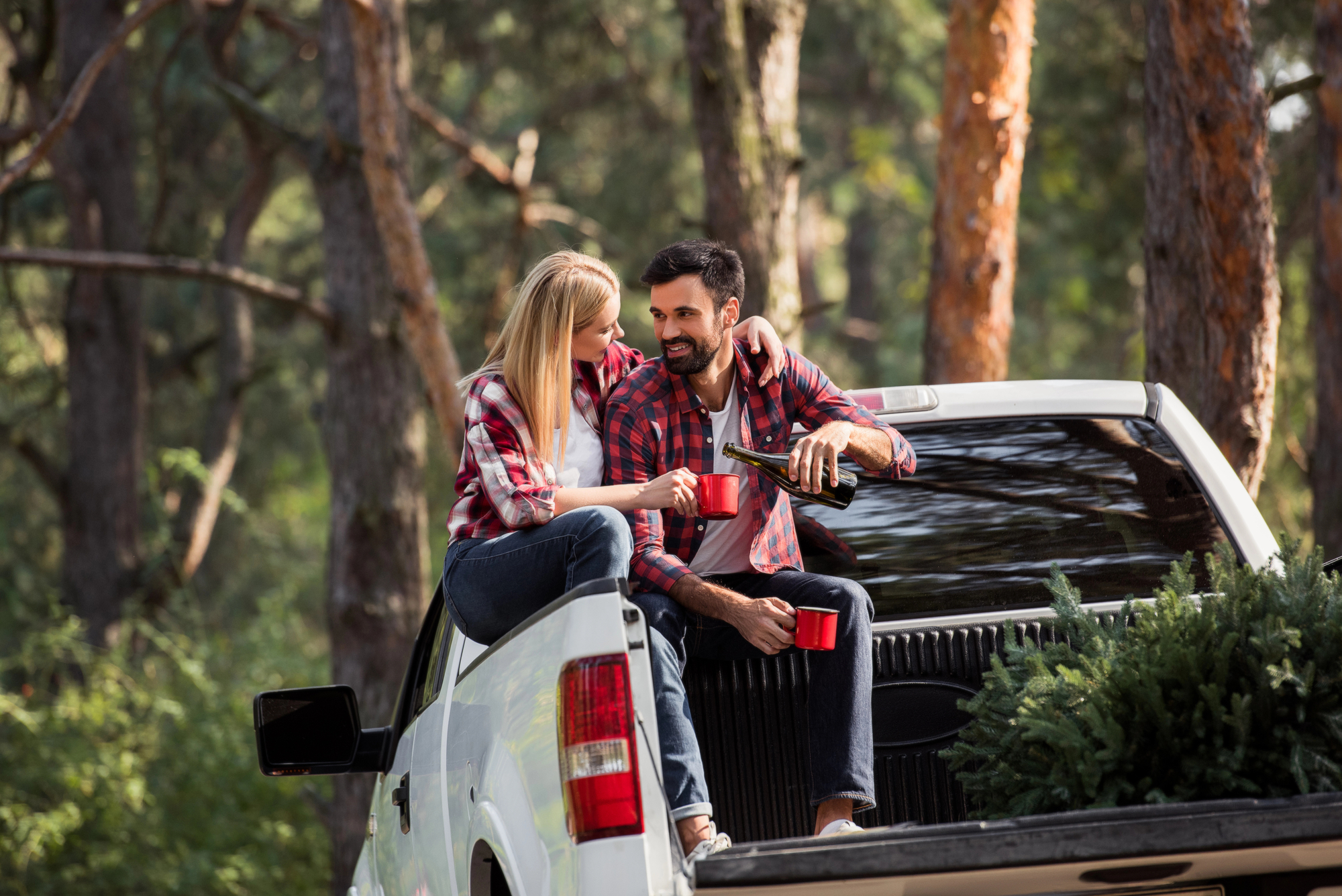 A couple sits on the open tailgate of a white pickup truck in a forest. They are smiling and holding red mugs, wearing casual, plaid shirts and jeans. A small Christmas tree is in the truck bed beside them. Trees and greenery surround them.