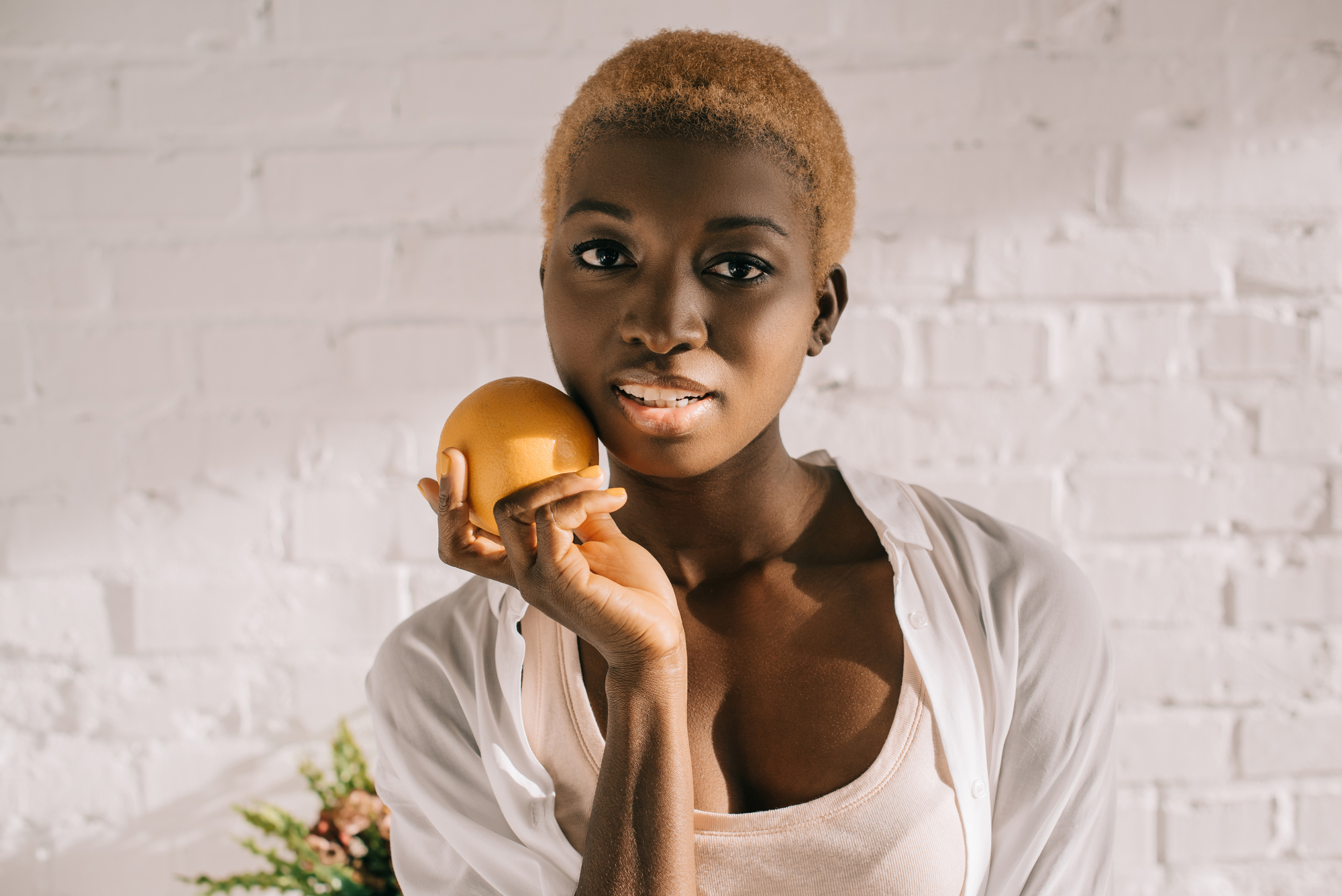 A person with short blond hair holds an orange close to their face, smiling slightly. Wearing a white shirt over a light pink top, they stand against a white brick wall in soft natural light. Some greenery is visible at the bottom left corner.