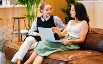 Two women sit on a brown leather couch in an open, modern office space. One is holding a laptop on her lap while gesturing as she speaks. The other woman listens attentively, sitting with one leg crossed over the other. Plants and barstools are visible in the background.