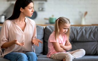 A woman with dark hair is sitting on a grey couch, speaking emotionally with her hands outstretched. Next to her, a young girl with blonde hair in braids sits with arms crossed and a pout, looking away. Both appear to be in a tense conversation.