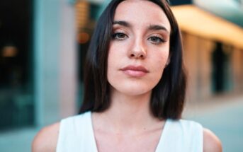 A woman with long dark hair and light skin stands outdoors, facing the camera. She is wearing a sleeveless white top, with her expression neutral. The background is slightly blurred, hinting at an urban setting with large windows and ambient lighting.
