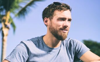 A man with a beard and sunglasses resting on his head sits outdoors against a clear blue sky. He wears a light blue t-shirt, and a palm tree is visible in the background, giving the scene a tropical feel. He appears to be looking off into the distance.