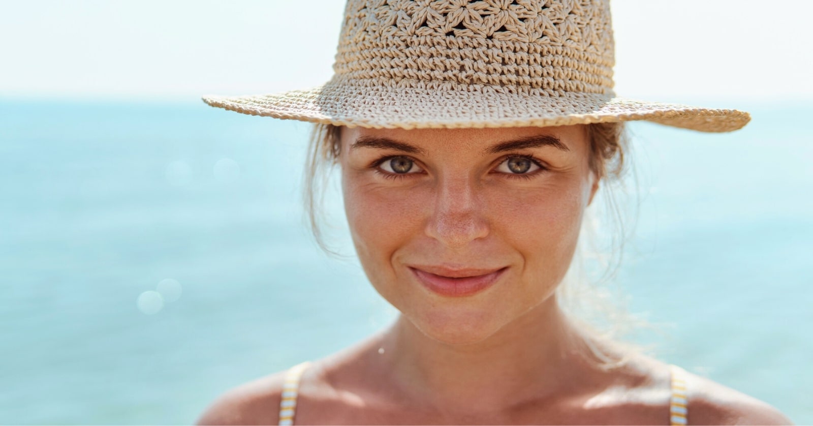Close-up of a person smiling at the camera, wearing a wide-brimmed straw hat. The background is blurred but appears to be a calm and sunny beach with blue ocean water. The person has a relaxed and happy expression on their face.