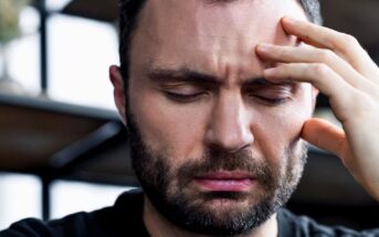 A close-up of a man with a beard, looking distressed or in pain. He has his eyes closed and is touching his forehead with his fingers, as if experiencing a headache. The background is blurred, suggesting an indoor setting with shelves.