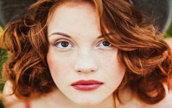 A person with fair skin, red wavy hair, and freckles looks directly at the camera with a neutral expression. They are wearing a dark hat and have light makeup, including red lipstick. The background is blurred greenery.