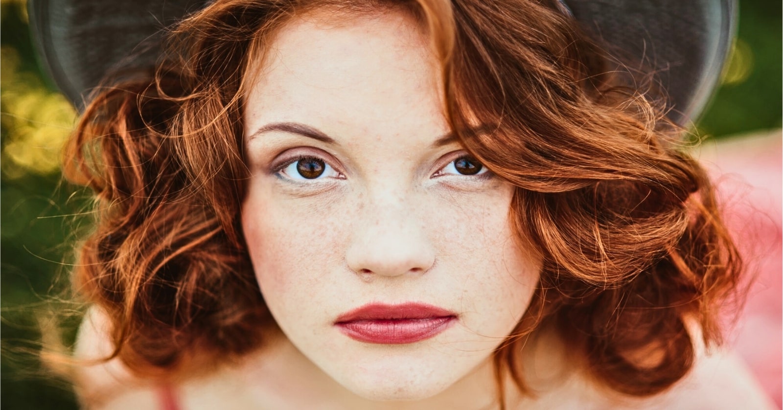 A person with fair skin, red wavy hair, and freckles looks directly at the camera with a neutral expression. They are wearing a dark hat and have light makeup, including red lipstick. The background is blurred greenery.