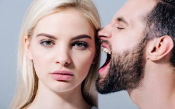 A woman with blonde hair and a neutral expression stands while a man with a beard next to her appears to be yelling or making a loud noise toward her ear. The background is plain and gray.