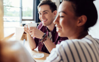 A young man and woman smile and enjoy a conversation at a bright cafe. The man holds a white coffee cup in both hands, while the woman looks at something out of the frame, smiling. They are seated at a wooden table with natural light coming in through large windows behind them.