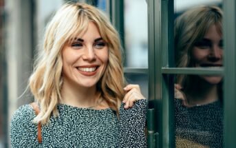 A woman with shoulder-length blonde hair smiles while standing by a window with a reflection of her visible in the glass. She is wearing a black and white patterned top and holds the edge of the window frame, exuding a cheerful and relaxed demeanor.