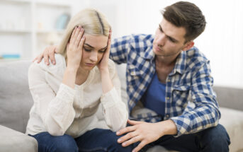A man in a blue plaid shirt sits beside a distressed woman with blond hair on a couch. He has his arm around her and his hand on her knee, offering comfort as she holds her head in her hands. They appear to be having a serious conversation.