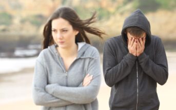 A woman in a grey hoodie stands with her arms crossed, looking away in frustration, while a man in a dark hoodie stands behind her with his head in his hands, appearing distressed. They are outdoors with a blurred beach and cliff background.
