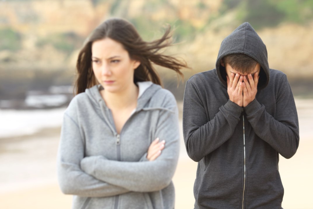 A woman in a grey hoodie stands with her arms crossed, looking away in frustration, while a man in a dark hoodie stands behind her with his head in his hands, appearing distressed. They are outdoors with a blurred beach and cliff background.