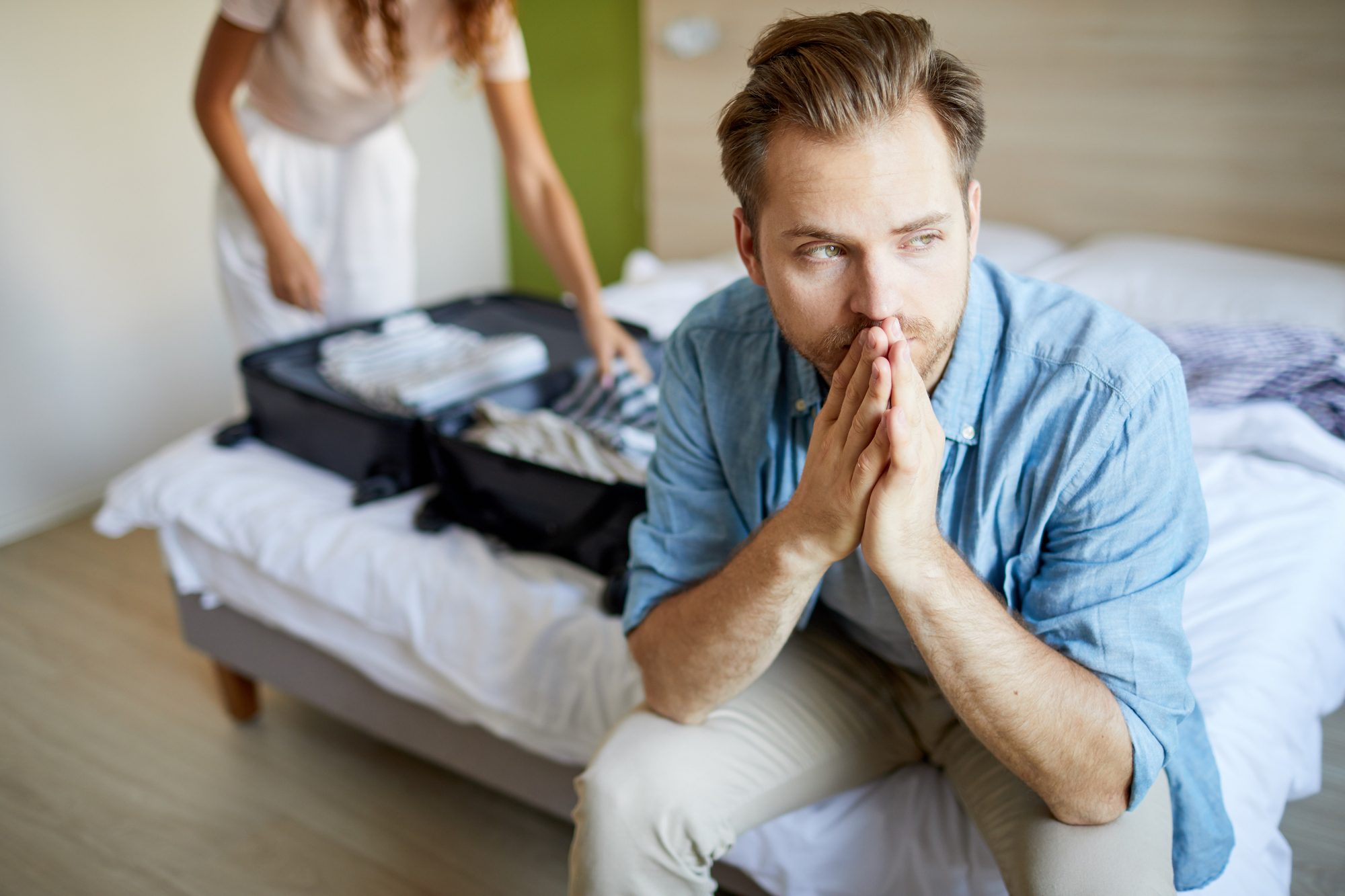 A man sits on the edge of a bed with his hands clasped near his mouth, looking pensive. In the background, a woman with curly hair is packing a suitcase on the bed. The room has light wooden flooring and a modern, minimalist design.