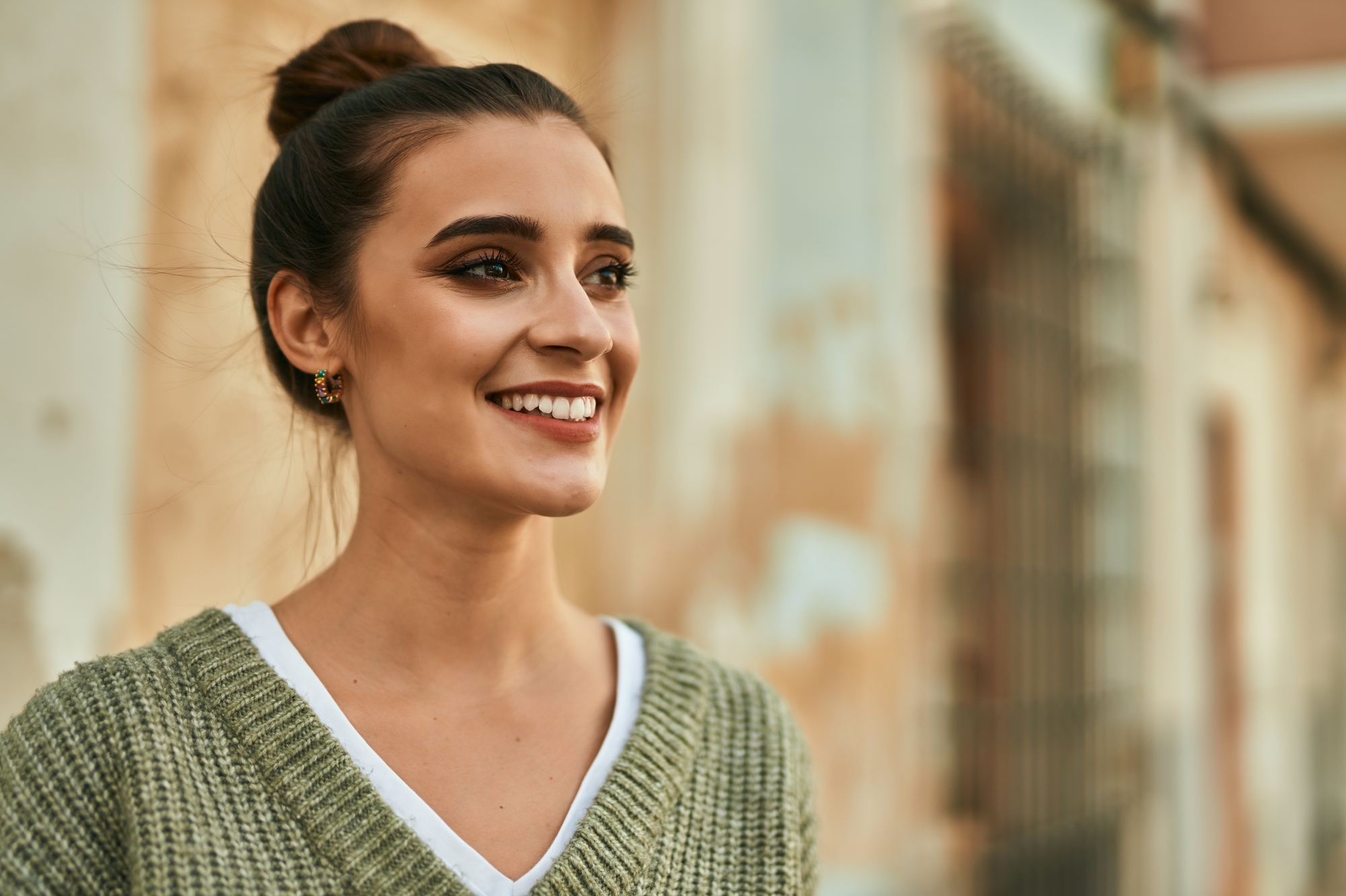 A young woman with dark hair tied in a bun smiles while looking to the side. She is wearing a green knit sweater over a white V-neck shirt. The background is blurry with warm-toned and pastel-colored walls.