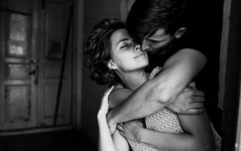 A black and white photo of a couple embracing. The woman, with short curly hair and closed eyes, leans back slightly against the man's chest, who is leaning in close and holding her tightly. They are standing in a dimly lit room with a slightly open door in the background.