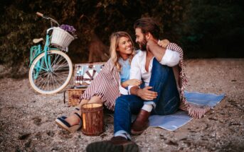 A couple sits on a beach blanket, smiling at each other. A bicycle with a flower-filled basket is parked nearby. They are surrounded by picnic items including a basket and small wooden bucket. The scene is relaxed and intimate with trees in the background.
