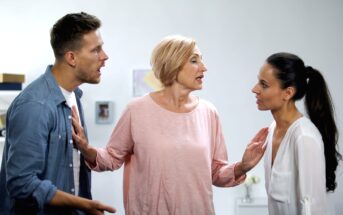 Three people are engaged in a heated discussion indoors. A man on the left and a woman on the right are facing each other, while an older woman in the middle is trying to mediate, extending her arms toward both of them. The atmosphere is tense.