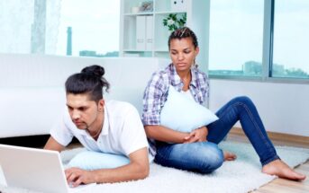 A man lying on a carpeted floor types on a laptop while a woman sits next to him, leaning against a couch and holding a pillow. They appear to be in a bright, modern living room with large windows in the background.