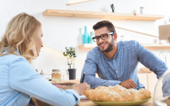 A man and a woman sit at a table in a cafe, engaged in a friendly conversation. The man, who is wearing glasses and a striped shirt, is smiling. The woman is writing in a notebook. In the foreground, there is a plate with pastries. The background shows shelves with decor.