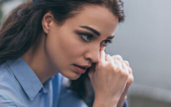 A close-up of a woman with long dark hair and a blue shirt. She looks pensive and concerned, resting her chin on her clasped hands while gazing into the distance, deep in thought. The background is blurred, emphasizing her expression and emotions.