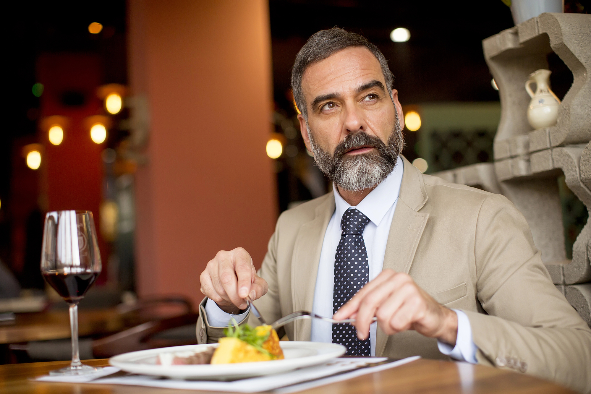 A distinguished man with a beard and mustache, wearing a beige suit and a navy polka-dot tie, is seated at a restaurant table. He is holding a fork and knife, about to eat from a plated meal. A glass of red wine is on the table beside him.